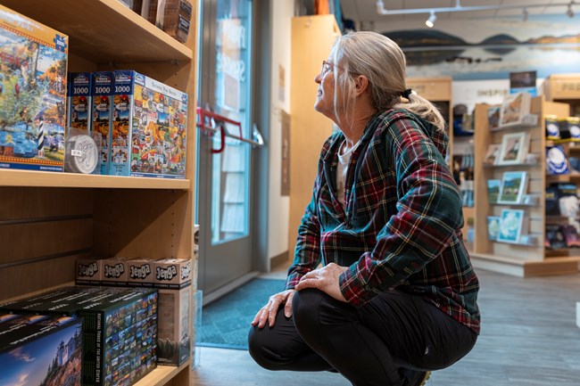 A visitor in a plaid shirt squats while browsing puzzles and games on a shelf in Acadia National Park's Park Store.