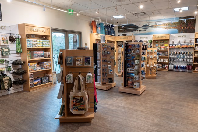 Interior of Acadia's Park Store with wooden shelves and racks displaying games, clothing, and souvenirs, under signs encouraging observation and preservat, and a lake mural in the background.
