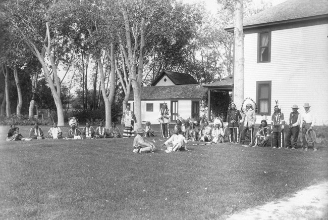 A meeting between James Cook and Jack Red Cloud on the lawn in front of the ranch house. Other people gather around in a semi-circle around them. Large trees stand in the background.
