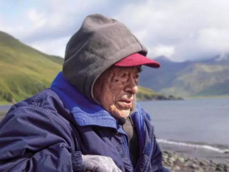 an old man wearing a hood over a red baseball cap looks out to the ocean with mountains in the distance.
