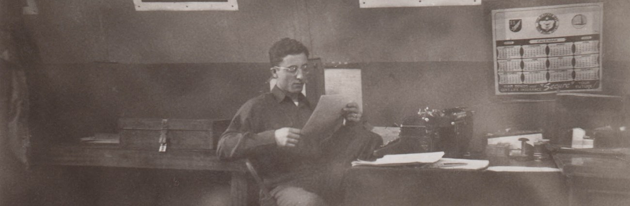 in a historic scene, a young man with glasses reads a paper sitting at a messy desk with war posters on the wall behind him.