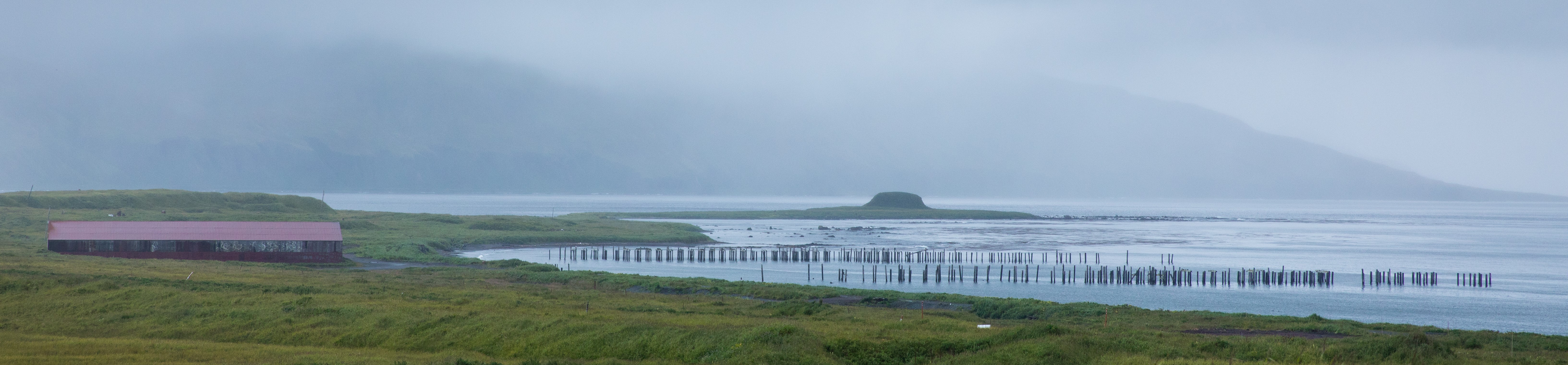 wooden pillars of an old, washed-away dock line the entrance to a coastal bay with a long, red building and grassy fields with foggy mountains in the background.