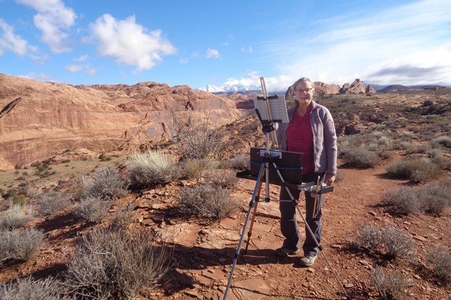 light-skinned woman painting at easel amidst desert shrubs, sandstone wall and distant mountains in background, blue sky with patchy clouds