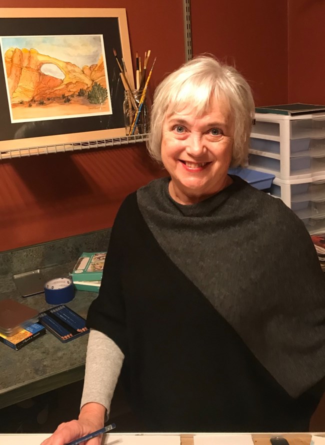 A woman wearing a black and grey sweater holding a pencil standing in her studio. A painting of a red rock landscape sits on a shelf in the background.
