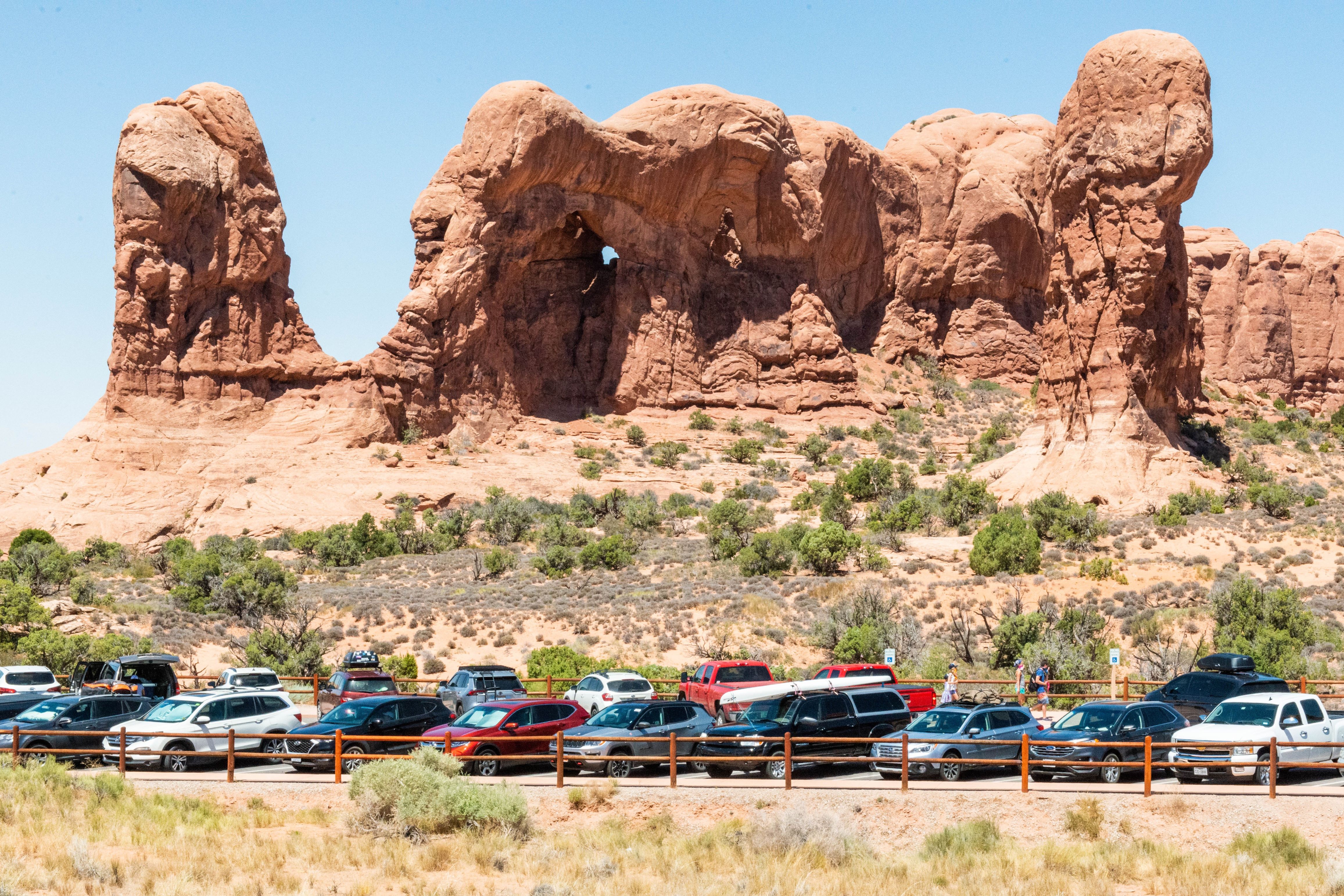 large stone columns and arches loom above vehicles parked along a fence, blue sky above