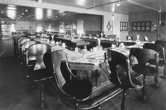 Black and white photograph of the lunch counter at Bright Angel Hotel in 1915. There are many empty chairs that are anchored into the ground, surrounding a rectangular lunch table, with napkins and silverware at every chair.