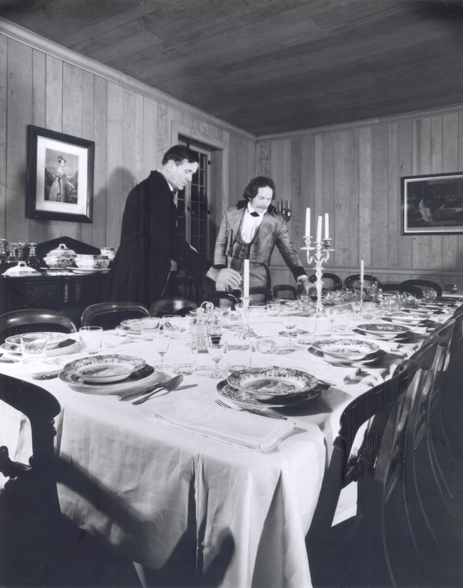 A black and white photo of two re-enactors setting the mess hall dining table inside the Chief Factor's House at Fort Vancouver.