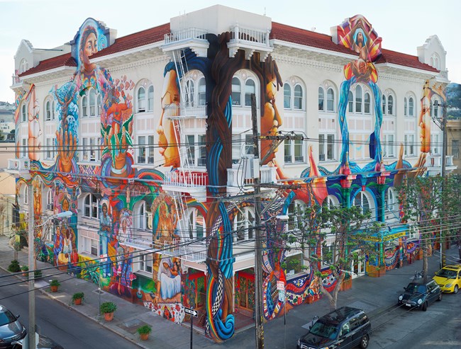 Corner view of four-story, symmetrical white stucco building covered with vibrant mural celebrating women from various cultural and ethnic groups and across time.