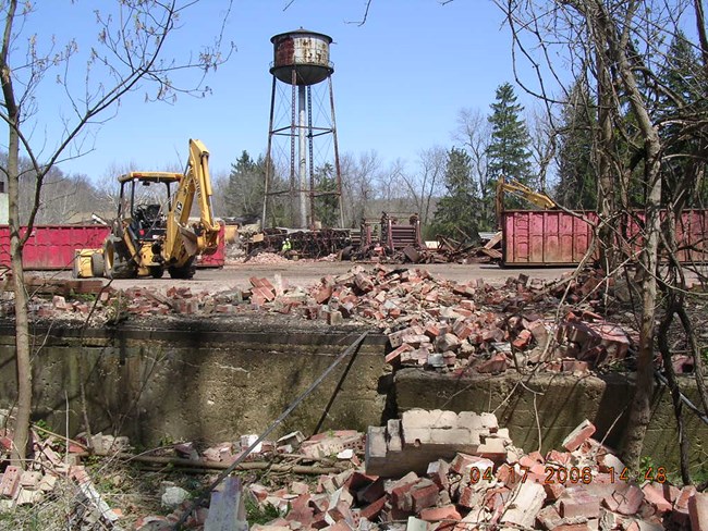 Below a rusting water tower, backhoes move stacks of bricks into red dumpsters.