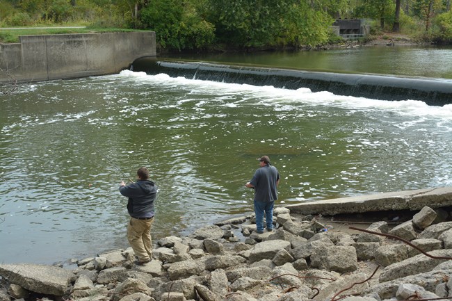 Two people fish from the near, rocky shore of the river; upstream of them water flows over the dam.