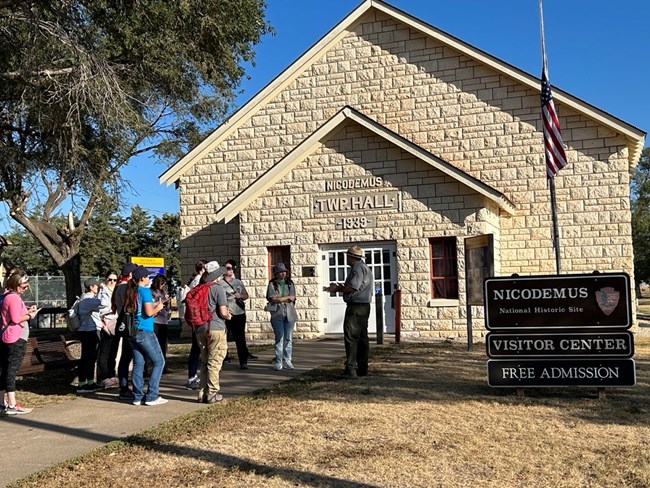 A white male park ranger talking to a group of people in front of an old limestone brick building, the Nicodemus Township Hall. In front of the building is a flagpole and brown sign that reads "Nicodemus National Historic Site".