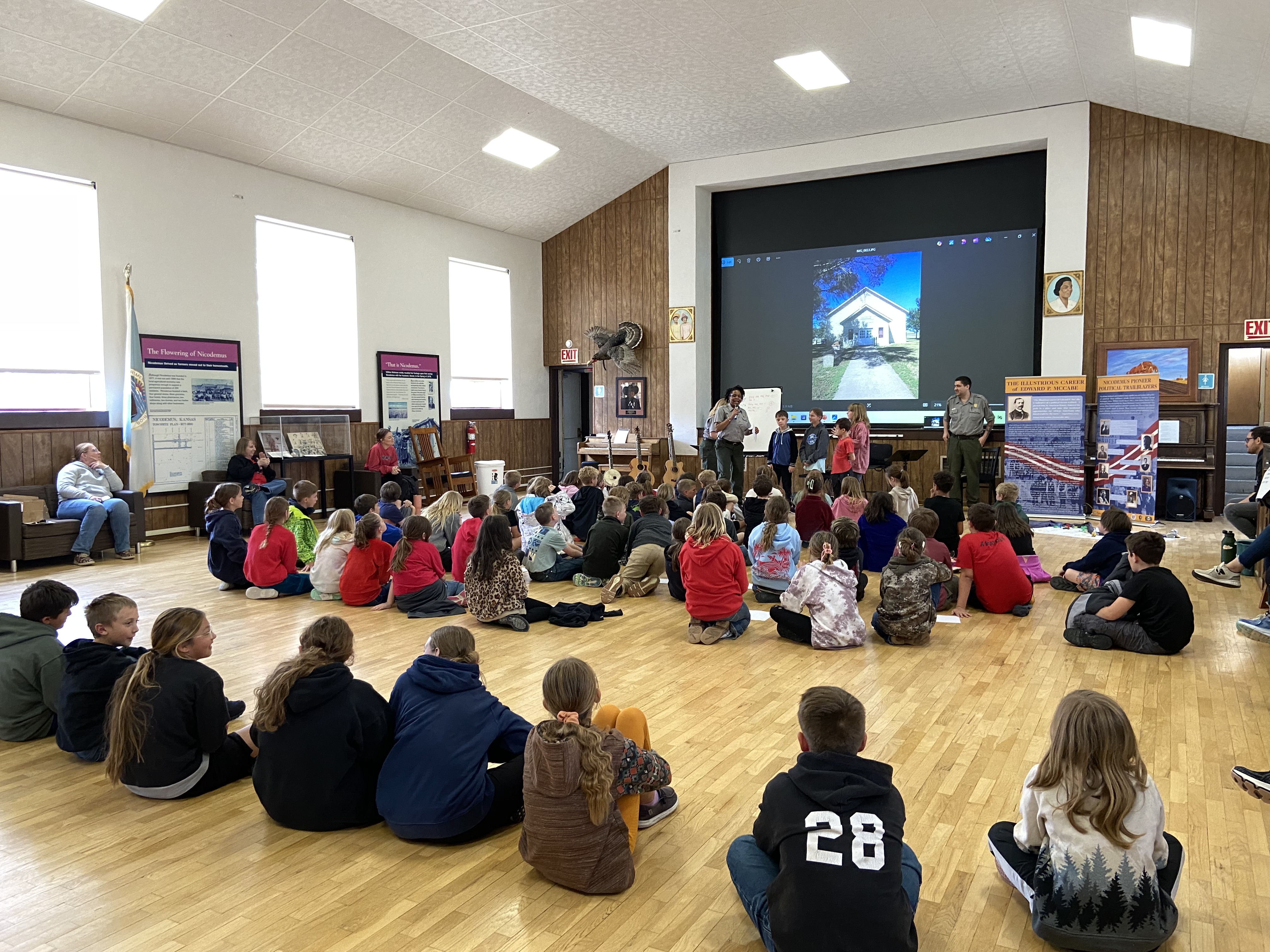 A large group of young students sit on the wooden floor of a large hall, their backs to the camera, and face a park ranger and four students standing in front of a stage and a projection screen showing the photo of a limestone building.