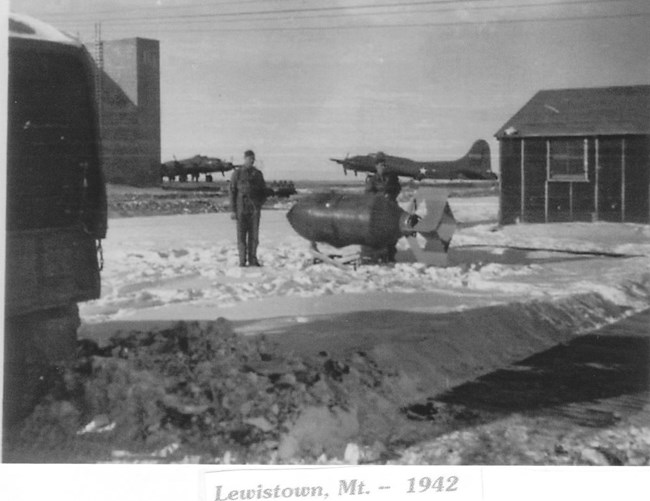 black and white photo of men in the snow next to a bomb. Bomber plane in background