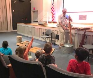 A man in a Volunteer-in-Parks uniform demonstrates playing an instrument in front of a group of children.