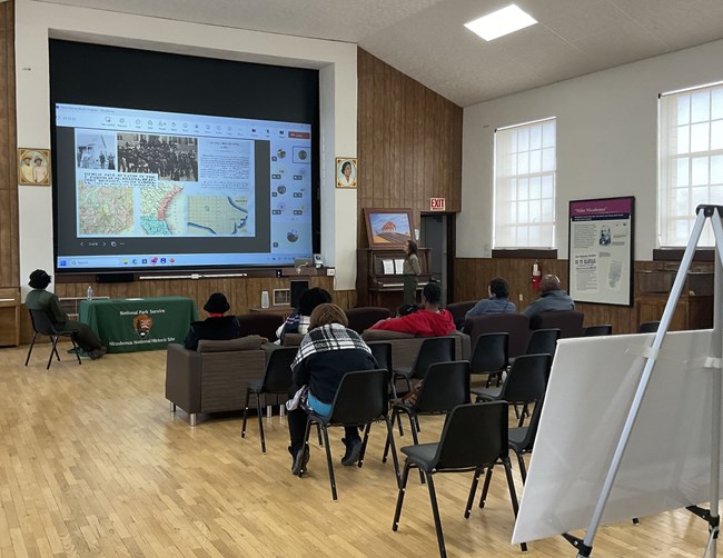 In a large hall, people sit in two rows of brown armchairs and two rows of black plastic chairs and face away from the camera, looking at the stage area, which is covered by a large projection screen showing a presentation with maps and images.