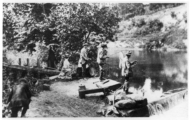 A black-and-white photo of men in coats and hats fishing from the shore above the dam; women sit and stand in long dresses.