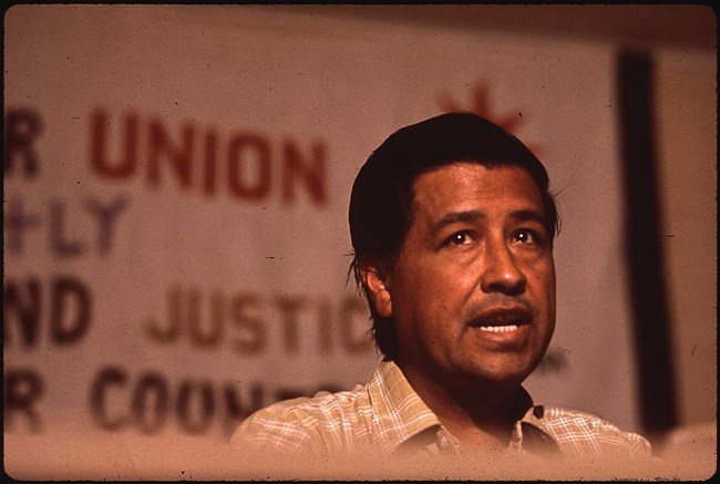 Photograph shows Cesar Chavez, photographed from below the table he is sitting at, in the middle of speaking. Behind him hangs a banner with words like “union” and “justice.”