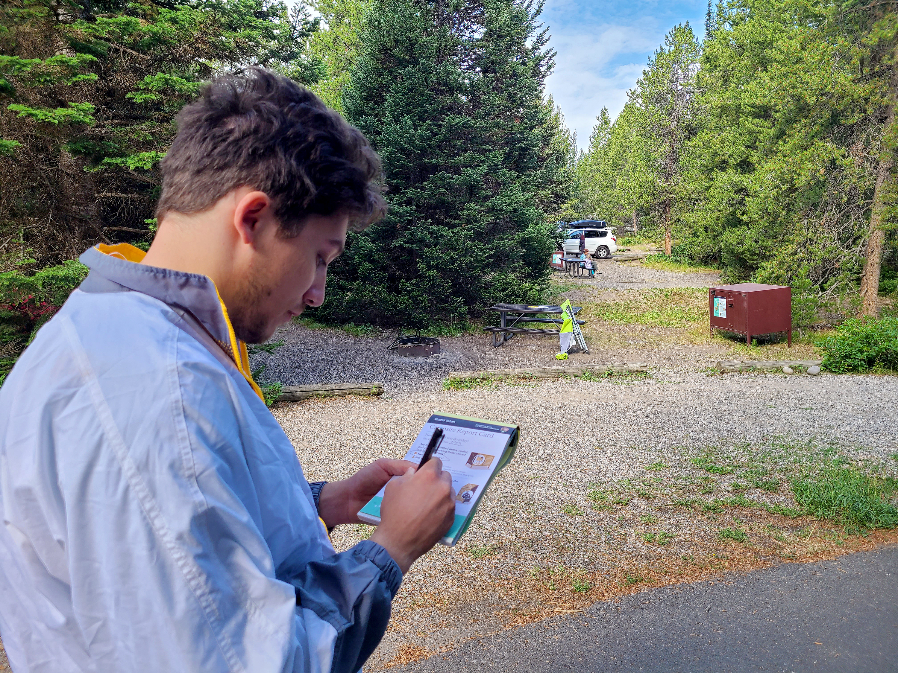 A man writing on a small form labeled "campsite report card." A campsite with bear box and evergreen trees is in the background.