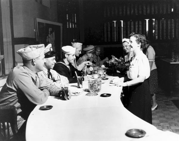 Black and white photo of a table. White servicemen in uniform sit on the left side of the table, white women in white shirts and dark skits stand on the right.