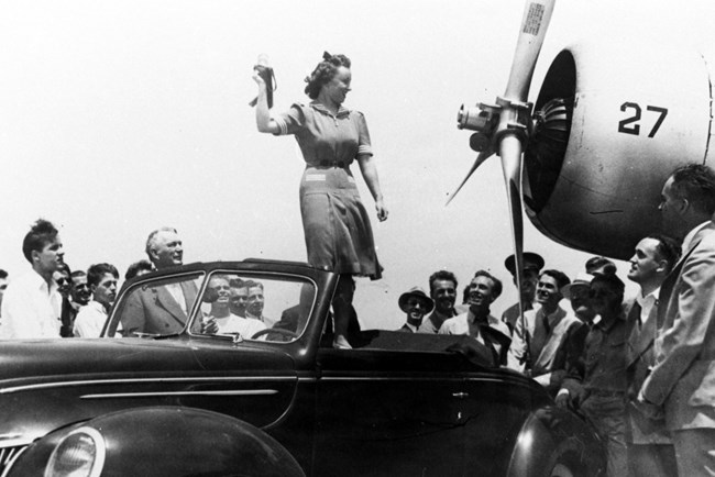 Black and white photo of a white woman standing on a car, arm up looking at a propeller of a plane.