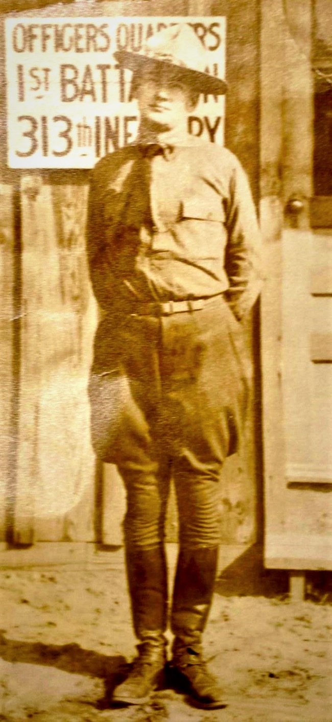 Man, standing, in military uniform, boots and hat, in front of a building, with a sign