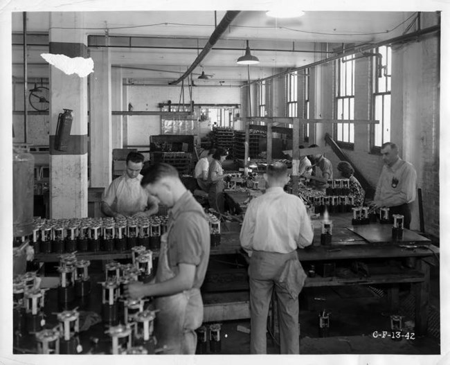 Black and white photo of white men with head bent over small cannisters on a conveyor belt