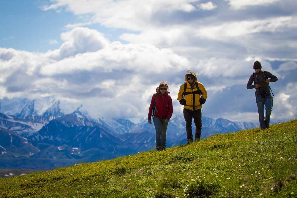 Three people hike on a tundra slope with the mountain range behind them.