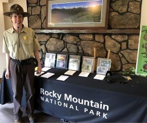 A young man in a Volunteer-in-Parks uniform stands in front of a Junior Ranger activity table.