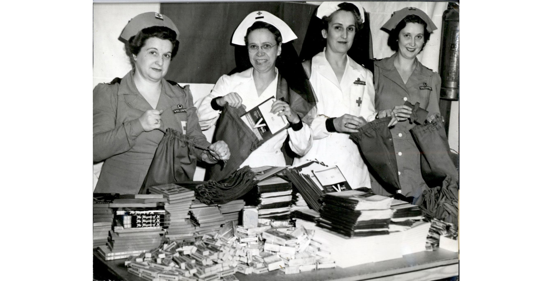 Black and white photo of 4 white women wearing white or tan Red Cross uniforms and nurses' caps. They are putting booklets and other items into bags from a table in front of them.