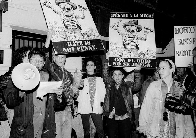 Group of women protestors. Individual on far left speaks into a bullhorn while reading off a page. Second and fourth from left hold large posters over their heads. Poster features cartoon skeleton with raised arms wearing fedora, suit jacket, and pants.