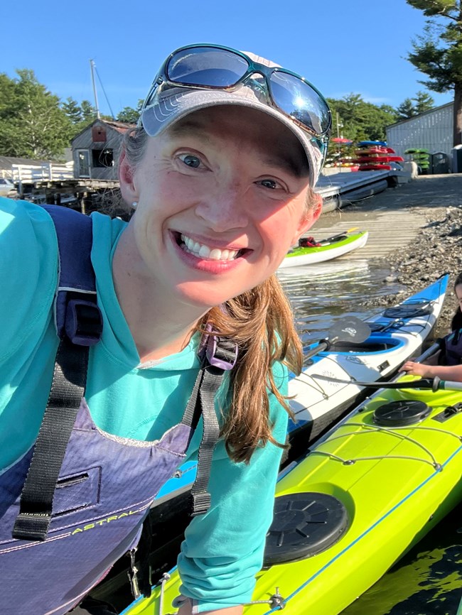 a smiling woman stands among kayaks on land