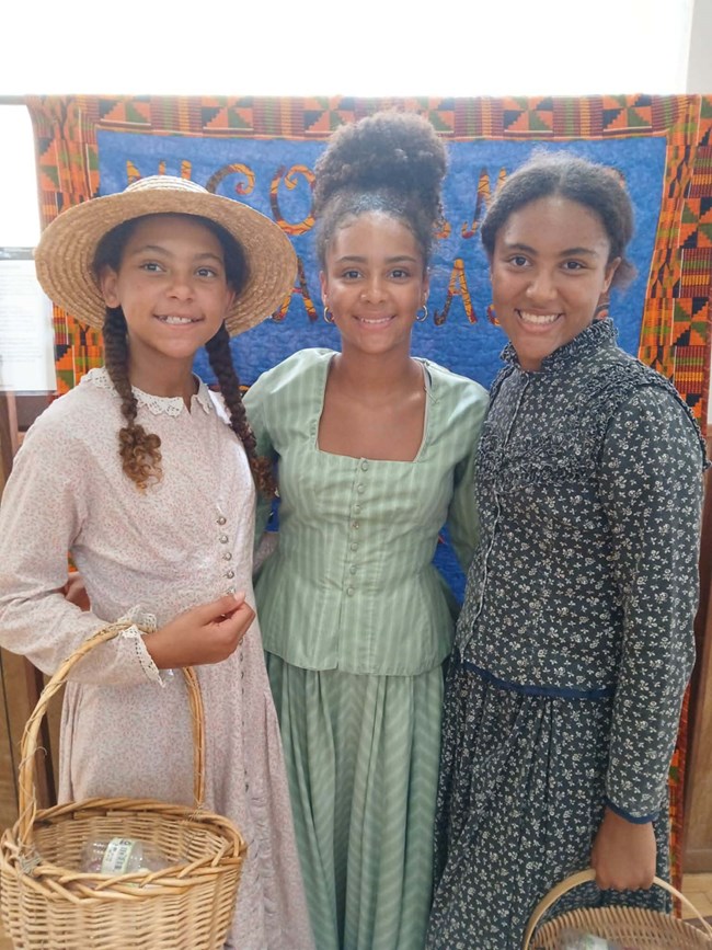 Three Black teenage girls, dressed in 1889 fashions, smile and pose in front of a bright blue and orange patterened quilt.