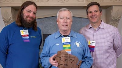 A man stands holding an award, on either side of him stands two men smiling. 