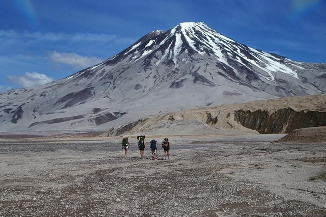 A group of four hikers walk toward a volcano.
