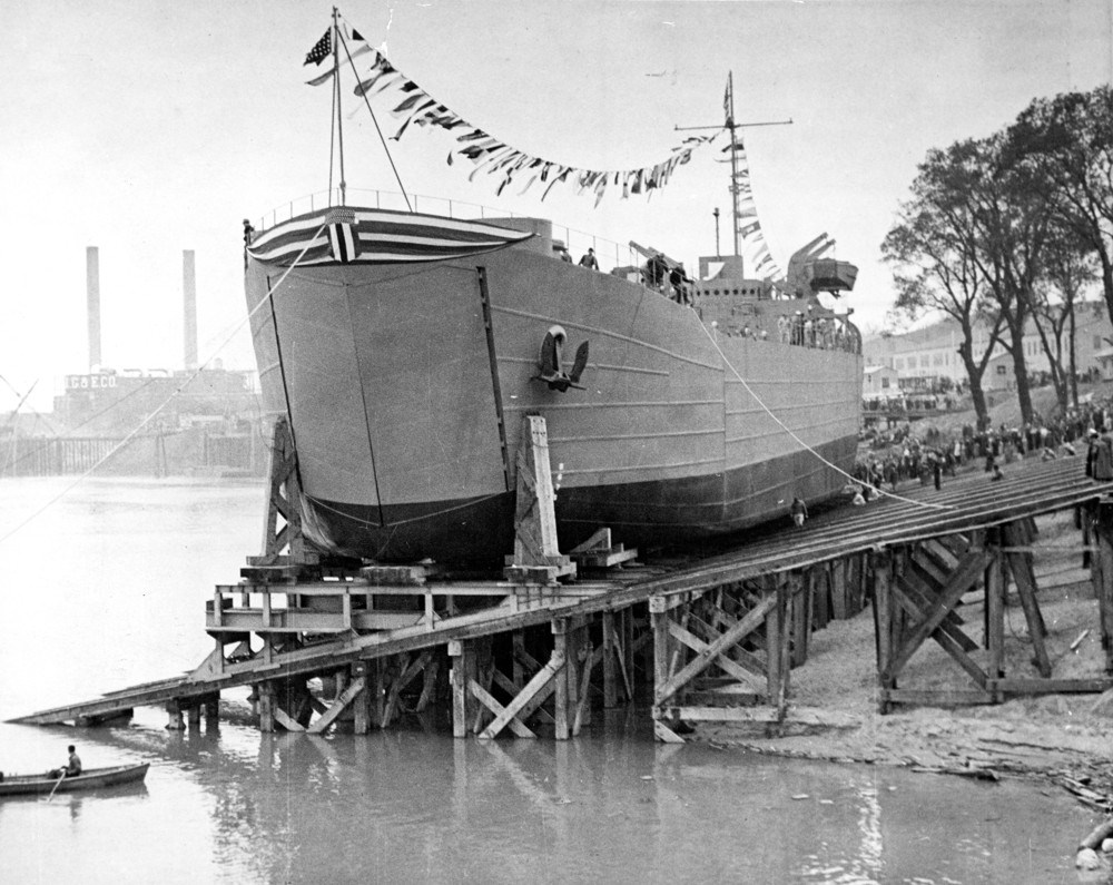 Black and white photo of a ship sitting on a wooden scaffolded dock. Flags fly between the mast and smokestacks are visible inbackground.