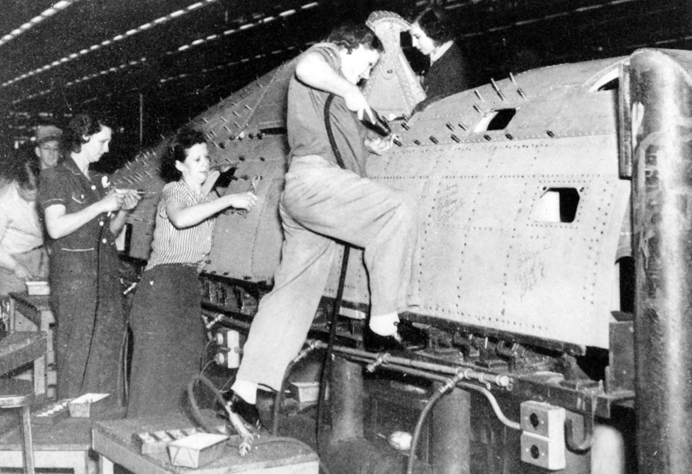 Black and white photo of women in work pants and coveralls drilling the rest of the plane.