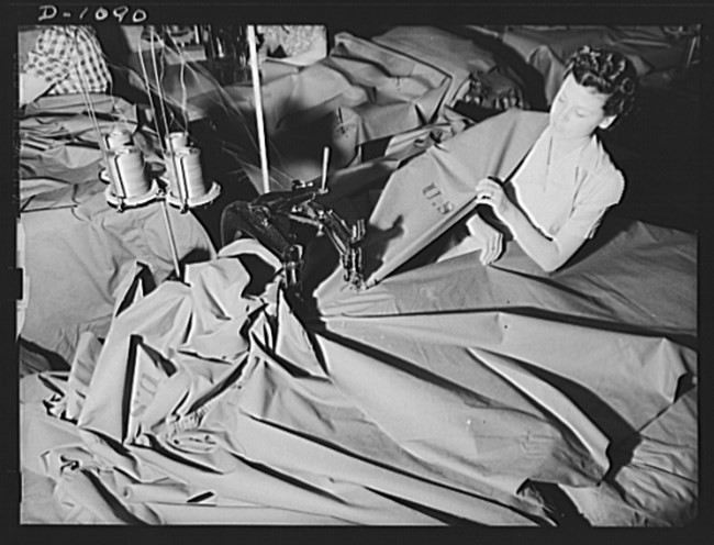 Black and white photo of a white woman at a sewing machine with a big piece of fabric. Other sewing stations visible in the right top corner.