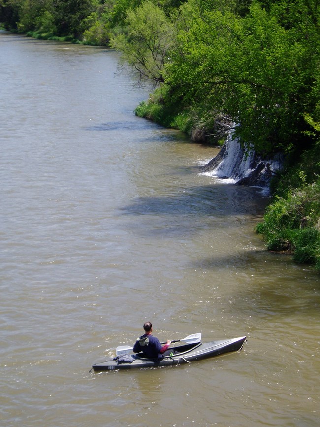 Kayaker on river, Waterfall along bank, blue sky above.
