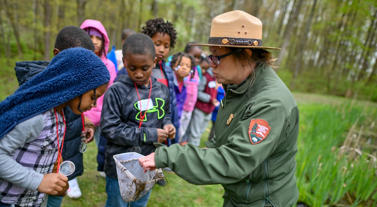 A woman in a green uniform jacket and brown hat with flat brim points to a net to children outside.