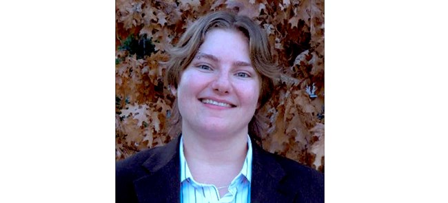 Headshot of woman with brown hair and dark jacket against a backdrop of brown leaves