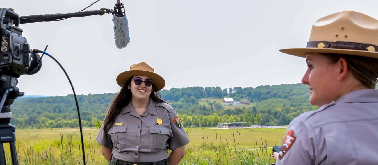 A woman in a gray uniform shirt a tan hat with a flat brim smiles in front of a camera outside.