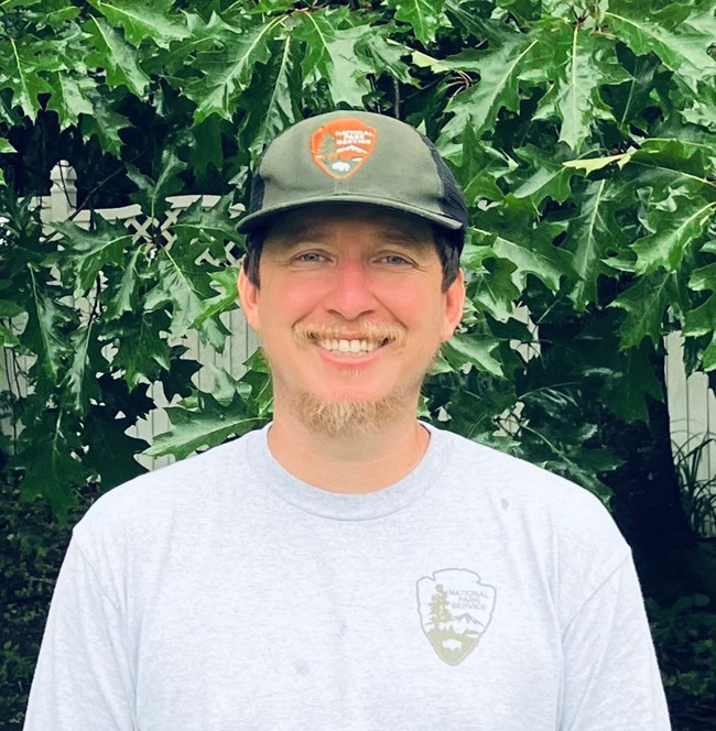 a man in a hat embossed with the National Park Service arrowhead smiles at the camera