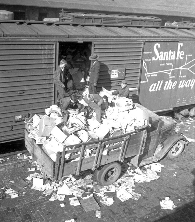 Black and white photo of a truck filled with newspaper piles onto a train