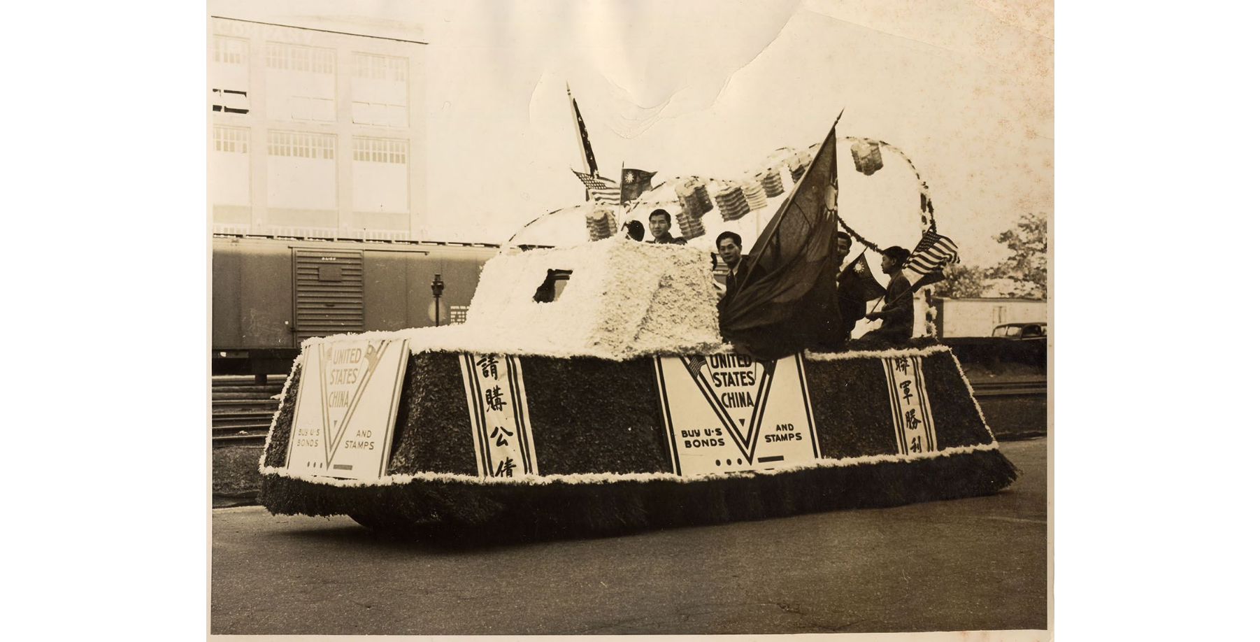 Black and white photo of a paper machier covered car. Flags are hanging from the top. A few men of Asian descent are visible sitting in the bed of the car.