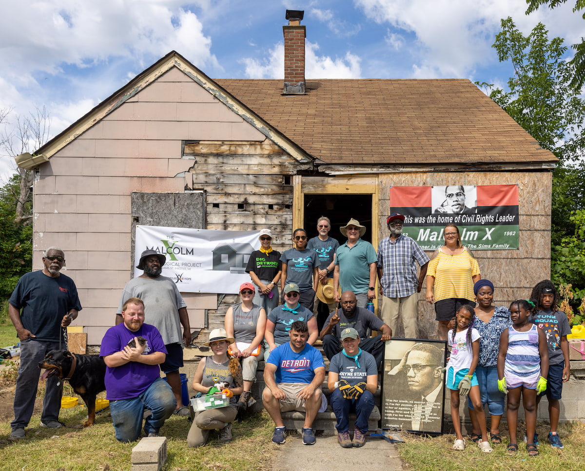  A group of 19 people stand in front of a house with two banners draped across the front. The house has particle board over the windows and one half of the house was stripped down to plywood.