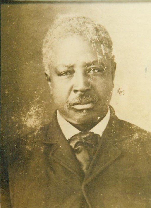 Sepia photo of a Black man from the shoulders up. He is wearing a black suit with white shirt and black tie, and has short white hair and short-trimmed facial hair.