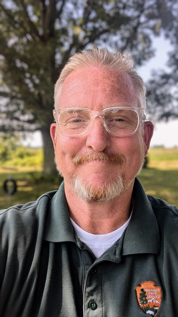 A smiling man with short blonde hair and a trimmed beard, wearing clear glasses and a dark green polo shirt with the National Park Service logo. He is standing outdoors with a tree and blurred greenery in the background, under daylight conditions.