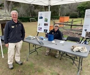 A man and a woman are pictured by a table filled with educational materials wearing Volunteer-in-Parks uniforms. 