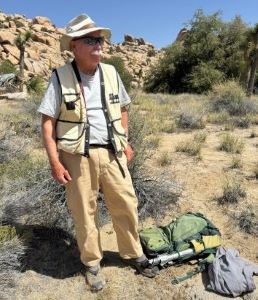 A man is shown standing with field gear in a desert environment.