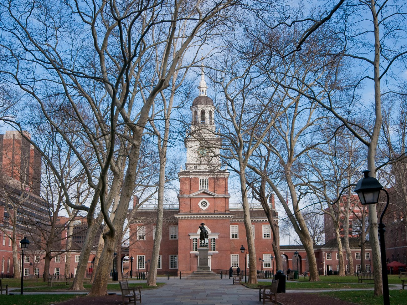Two story brick building with white clock tower and cupola and a statue of a man in a tricorner hat standing on a pedestal